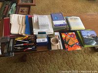 Wide view of large assortment of office supplies including notebooks, binders, pens, pencils, paper packs, and other desk items laid out on a table.