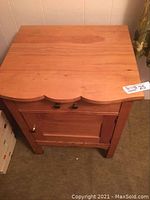 Front view of a pine side table showing a scalloped edge top, drawer with two knobs, and cupboard door with a knob.
