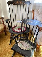 Pair of two black painted wooden rocking chairs, larger and smaller size, each with floral detailing on the chair back and vertically arranged spindles in the backrest. Custom braided fabric cushions are on each seat.
