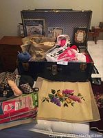 Wide shot of the large vintage trunk open with various embroidered pieces, yarn, and books visible inside and in front of it on a flat surface.