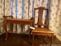 Photo showing the wooden side table with curved legs and wicker top next to the wooden chair, positioned against a curtain with leaf pattern.