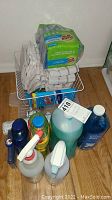 Photo showing various bottles of cleaning liquids, spray bottles, dish towels, wire dish rack and sponges arranged on wooden floor.