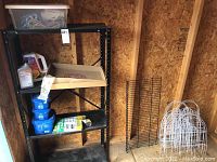 Wide view of black metal shelving unit holding gardening supplies with white metal fencing leaning against shed wall