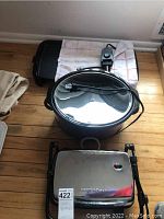 View of crock pot with glass lid, Hamilton Beach panini press with stainless steel lid, and flat electric griddle all placed on a wooden floor with a towel beneath the griddle.