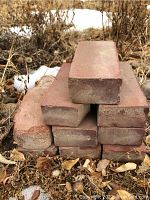 Stack of reddish rectangular landscaping bricks placed outdoors on ground with dried leaves and sparse snow around.