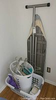Corner with two laundry baskets stacked, ironing board leaned against the wall with hangers visible inside basket