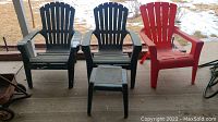 Three plastic outdoor chairs (two green Adirondack style and one red) with a small green plastic table on a porch with some snow on the ground in the background.