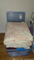 View of folded quilts and blankets stacked in a blue plastic storage tub, showing the top quilt with pink and white floral/leaf pattern.