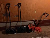 Two snow shovels with blue handles, Toro electric snow shovel S-120 in red and black, and orange extension cord coiled beside it on garage floor.