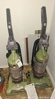 Two green Hoover WindTunnel bagless upright vacuum cleaners standing next to each other with visible dust canisters and upright handles