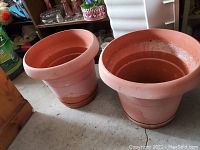 Two large terracotta planters on a concrete floor indoors near shelves with miscellaneous items.