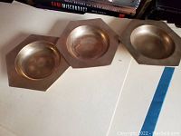 Three metal trays on a beige surface, two with hexagonal bases and one round tray all having slightly raised edges.