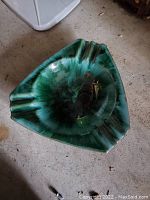 Photo of a triangular green glazed pottery bowl with notches, used as an ashtray, placed on a concrete floor.