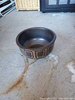 Photo of a dark brown round ceramic bowl with rectangular and square patterns around the outside, sitting on a concrete floor.