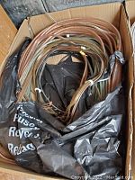 Top-down photo showing a cardboard box filled with several coiled lengths of insulated electrical wires in brown, black, and green, with some wires tied together and a black plastic bag protecting parts of the contents.