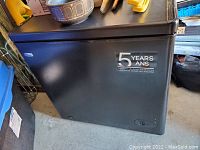 Photo of a black deep freezer with a 5-year guarantee sticker on front, set in a cluttered storage area.