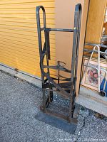 Full view of the heavy duty black metal hand truck dolly standing upright on pavement with a yellow garage door in the background.
