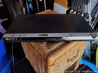 Black LG DVD player placed on wicker basket, showing front and top surfaces in a indoor setting.