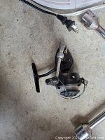 Photo of a black and silver fishing reel with attached handle and fishing line, placed on a concrete surface with metal objects nearby.