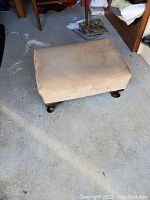 Foot stool covered in beige fabric with wooden legs on a concrete floor.