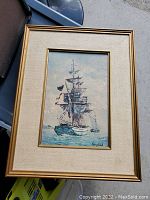 Framed painting of a sailing ship with multiple sails and flags signed by Keirstead in gold frame with beige fabric mat.