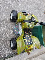 Green kids ATV toy with black seat and handlebars, decorated with colorful stickers, photographed lying on pavement showing top and side view.