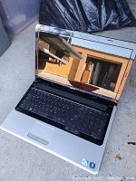 Black Dell laptop open showing keyboard and reflective screen with Windows 7 sticker on palm rest, photographed on concrete surface.