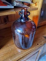 Photo of a large brown glass jug placed on a wooden surface showing front and side views of the jug details.