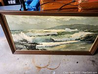 Framed seascape painting showing crashing ocean waves and coastline under a cloudy sky, visible signature at bottom right