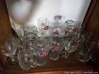 Group of 16 clear glass beer mugs and beer glasses showing various beer logos placed on shelf.