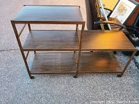 Photo of metal shelving unit with two connected sections, featuring dark metal frame and three brown wood-look shelves on casters, placed outside on pavement near chairs and framed art.