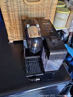 Black Nespresso coffee machine by DeLonghi with chrome lever and side milk frother container on black surface, showing front and side view.