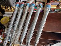 Six metal ground anchors with spiral screw design packaged with labels showing outdoor usage instructions and imagery