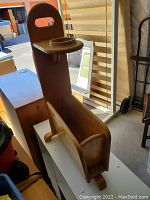 Wooden child high chair with backrest, front tray, and open storage box beneath the seat, placed indoors on a white surface among other items.