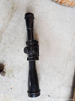 Black rifle scope with adjustment knobs, shown lying on a concrete floor.