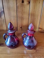 Two red glass containers with rounded bodies, handles, spouts, and pointed stoppers on a wooden surface