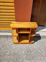 Photo of a small wooden shelving unit with curved side shelves taken outdoors on pavement next to wooden doors and a wall.