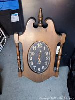 Photo shows full frontal view of wooden wall clock with decorative top finial and spindles on sides, black oval dial with gold numbers 1-12 and ornate hands.