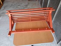 Wooden orange wall hanging shelf seen from above on a table, showing horizontal slats and pegs for hanging.