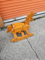 Wooden rocking horse made of light brown polished wood, placed outdoors in front of an orange metal garage door.