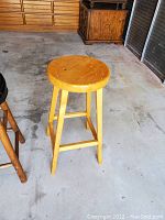 Wooden stool with light wood finish, round seat, and four legs, photographed in a garage-like setting on concrete floor.