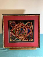 Full view of the framed mola textile artwork showing the colorful central face within a star pattern and surrounding geometric designs on black a fabric background, with red mat and dark wooden frame.