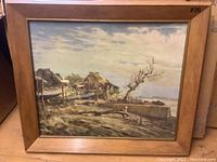 Frontal view of the framed painting showing a coastal scene with seaside shacks, a barren tree, and cloudy sky.