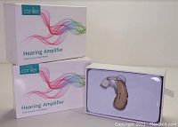 Photo of two Conlier hearing amplifier boxes stacked, and one open box displaying a beige hearing amplifier unit in molded plastic.