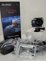 Photo showing Akaso V50 Elite action camera mounted on a white box next to the boxed packaging and an assortment of sealed accessories including waterproof case, charger, mounts, and remote.