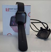 Front view of black smart watch with rectangular display on black strap, displayed in front of the original box. Charging cable with magnetic connector placed nearby.