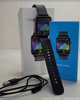 Photo showing the smartwatch fitted on black strap standing upright near its retail box and user manual with charging cable.