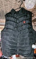 Black quilted heated vest hanging on hanger showing front zipper and collar.