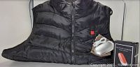 Black insulated heated vest size XXL by Genovega laid flat with visible front zipper and chest logo, along with a silver oval power pack/hand warmer device and its box on the right.