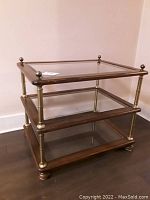 Full view of the 3 tiered side table showing wood frames with glass shelves and brass rods with decorative finials.